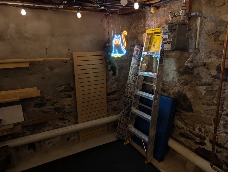 Basement corner with a neon cat sign, a stepladder, and daybed slats leaning against a stone wall